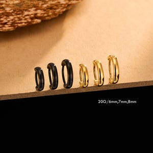 May include: A collection of six hoop earrings in varying sizes and colors. Three are black, and three are gold. The text "20G / 6mm, 7mm, 8mm" is printed below the earrings. The earrings are displayed on a brown surface.