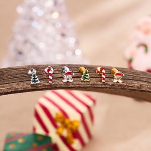 以下が含まれることがあります： クリスマスをテーマにしたスタッドピアスのコレクション。ピアスは、クリスマスツリー、キャンディケイン、サンタクロースのデザインが特徴です。木製の表面に表示され、クリスマスの装飾がぼやけた背景になっています。