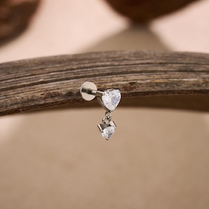 May include: A silver-colored lip piercing with two clear, heart-shaped cubic zirconia stones. The top heart is fixed, while the bottom heart dangles. The jewelry is set against a neutral background.