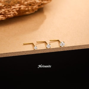 Pode incluir: Três piercings de nariz em forma de L em metal dourado, cada um com uma pedra Moissanite redonda e transparente numa cravação de quatro garras. Os piercings estão alinhados, com a palavra Moissanite escrita em branco por baixo.