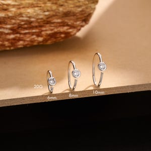 Op de afbeelding: Drie zilveren neusringen met heldere, ronde edelstenen. De ringen worden in verschillende maten getoond: 6 mm, 8 mm en 10 mm. De dikte is 20G. De sieraden staan tegen een neutrale achtergrond.