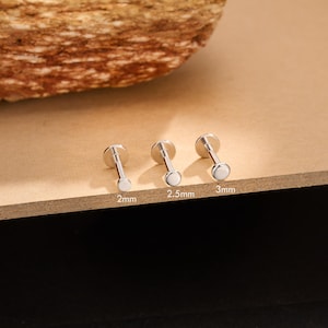 Puede incluir: Tres pendientes plateados con parte trasera plana y tapas circulares blancas. Los pendientes están etiquetados con los tamaños: 2 mm, 2,5 mm y 3 mm. Los pendientes se muestran sobre una superficie marrón claro.