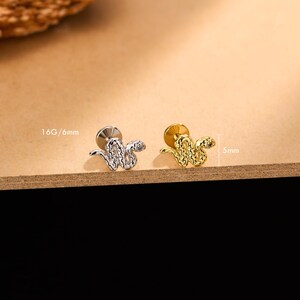 Pode incluir: Dois brincos em forma de cobra, um prateado e outro dourado, são exibidos em uma superfície de madeira. O brinco prateado está rotulado como 16G/6mm. O brinco dourado está rotulado como 5mm. Os brincos apresentam desenhos detalhados de cobras.