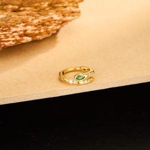 May include: A gold-colored snake-shaped ring with a green gemstone eye and small white gemstones. The ring is on a light brown surface with a textured, brown object in the background. Jewelry.
