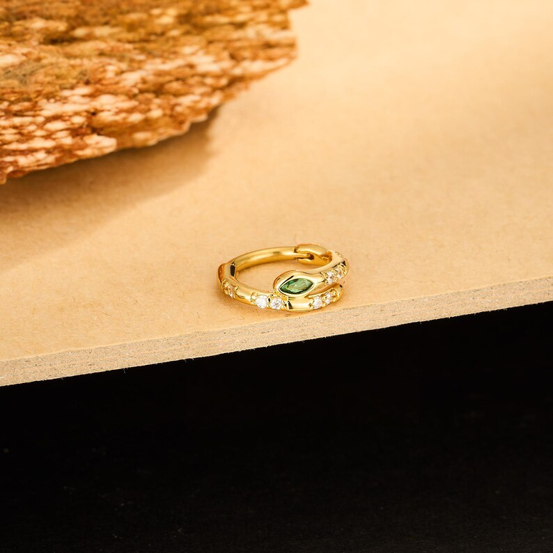 May include: A gold-colored snake-shaped ring with a green gemstone eye and small white gemstones. The ring is on a light brown surface with a textured, brown object in the background. Jewelry.