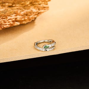 May include: A silver-colored hoop earring in the shape of a snake. The snake's head features a green gemstone eye, and the body is encrusted with small, clear gemstones. The earring is displayed on a light brown surface.
