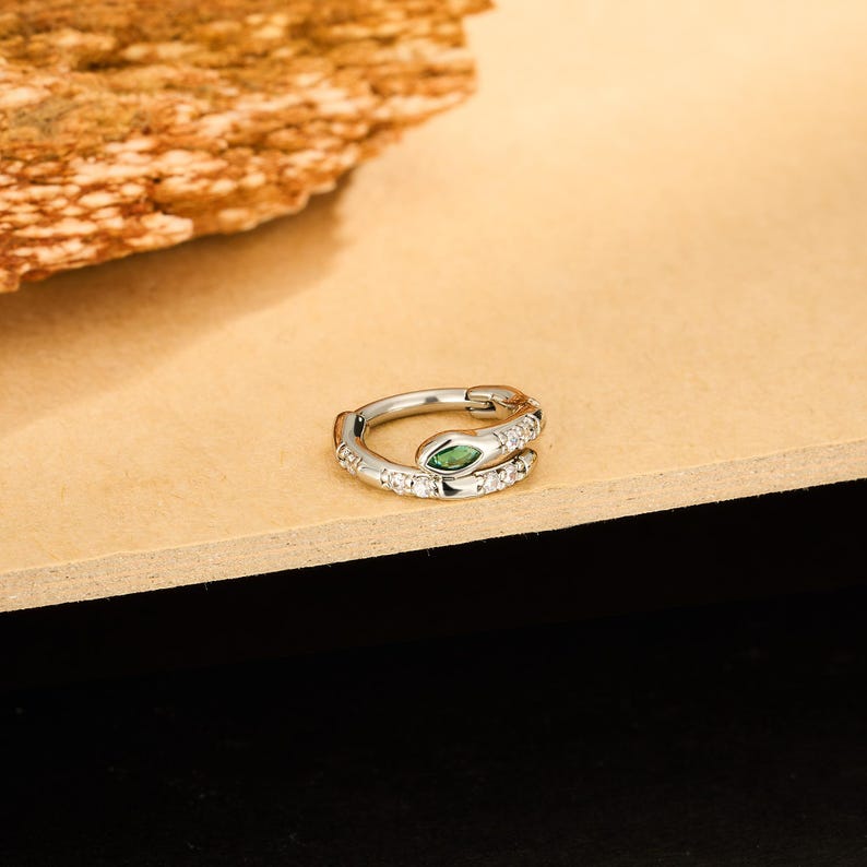 May include: A silver-colored hoop earring in the shape of a snake. The snake's head features a green gemstone eye, and the body is encrusted with small, clear gemstones. The earring is displayed on a light brown surface.