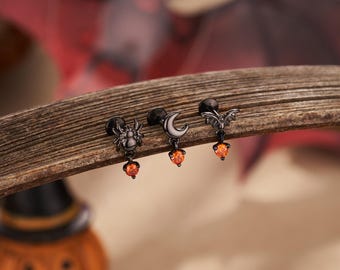 Op de afbeelding: Drie zwarte Halloween-thema oorbellen met oranje edelsteen accenten. De oorbellen hebben een spinnen-, halve maan- en vleermuisontwerp. Ze worden getoond op een stuk hout, met een vage Halloween-achtergrond.