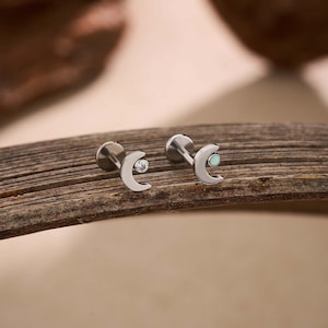 May include: Two silver-toned moon-shaped earrings with a small, round gemstone at the center of the crescent. One earring has a clear gemstone, and the other has a light blue gemstone. The earrings are displayed on a wooden surface.