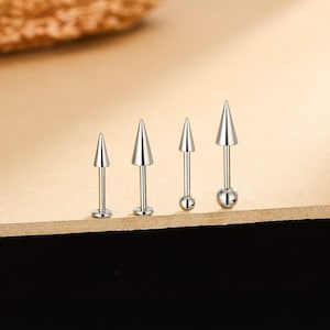 May include: Four silver-coloured, pointed stud earrings. Each earring has a conical spike design. The earrings are displayed on a light brown surface, with a dark brown background.