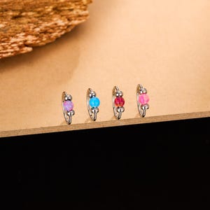 May include: Four small silver hoop earrings, each with a different coloured bead: purple, blue, red, and pink. The earrings are displayed on a light brown surface with a dark brown and black background.