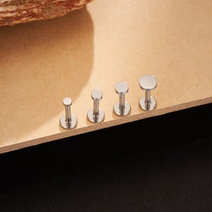 May include: Four silver-colored, flat-topped labret studs of varying sizes are arranged in a row. The studs have a post and a flat, circular top. The smallest stud is on the left, with the sizes increasing to the right.