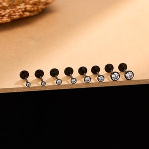 Könnte beinhalten: Eine Sammlung schwarzer Labret-Ohrstecker mit klaren, runden Zirkonia-Steinen. Der Schmuck wird auf einer hellbraunen Oberfläche präsentiert. Die Ohrstecker variieren in der Größe, wobei die Steine im Durchmesser zunehmen.