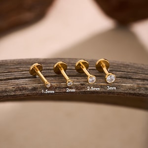 May include: Four gold-colored, flat-back earrings with clear, round gemstones. The earrings are labeled with sizes: 1.5mm, 2mm, 2.5mm, and 3mm. The earrings are displayed on a wooden surface.