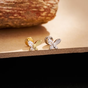 Puede incluir: Dos pendientes con forma de mariposa. Uno es dorado, el otro plateado, cada uno con una piedra central transparente en forma de lágrima y una piedra redonda más pequeña debajo. Los pendientes se muestran sobre una superficie marrón.