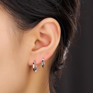 May include: A close-up of a person's ear with two small, silver hoop earrings. The earrings are square-shaped and have a polished, reflective surface. The background is dark, emphasizing the earrings and the ear's details.
