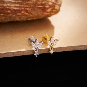 May include: Two small, decorative earrings. One is silver-toned, the other gold-toned. Each features a cluster of clear, marquise-cut stones forming a floral design, with a dangling, smaller stone. The earrings are displayed on a light brown surface.
