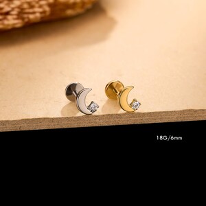 May include: Two flat-back stud earrings featuring a crescent moon shape with a small clear gemstone accent. One earring is silver-toned and the other is gold-toned. The text 18G/6mm is printed on the image, indicating the gauge and post length.