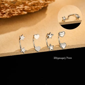 May include: Silver-colored curved eyebrow rings with star and heart designs. The jewelry is shown in a group of three, with a close-up of one ring. The text "20(gauge)/7mm" is visible.