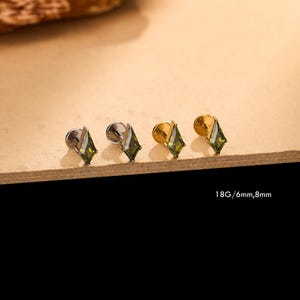 May include: Four labret stud earrings featuring kite-shaped olive green gemstones set in metal. Two studs have a silver-tone finish and two have a gold-tone finish. The text 18G/6mm,8mm is printed in the lower right corner of the image.