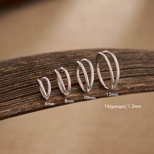 May include: A set of silver-colored double hoop earrings in various sizes: 6mm, 8mm, 10mm, and 12mm. The earrings are displayed on a wooden surface, with the size of each earring clearly labeled in millimeters and the gauge size.