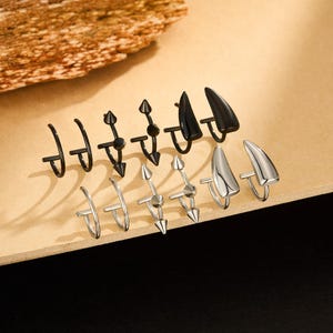 May include: An assortment of ear cuffs in black and silver tones. Designs include curved bars, spiked accents, and horn-shaped tips. The jewelry is displayed on a light brown surface.