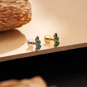 May include: Two gold-colored leaf-shaped earrings with three green gemstones each. The earrings have a flat back and a post for piercing. The earrings are displayed on a light brown surface.