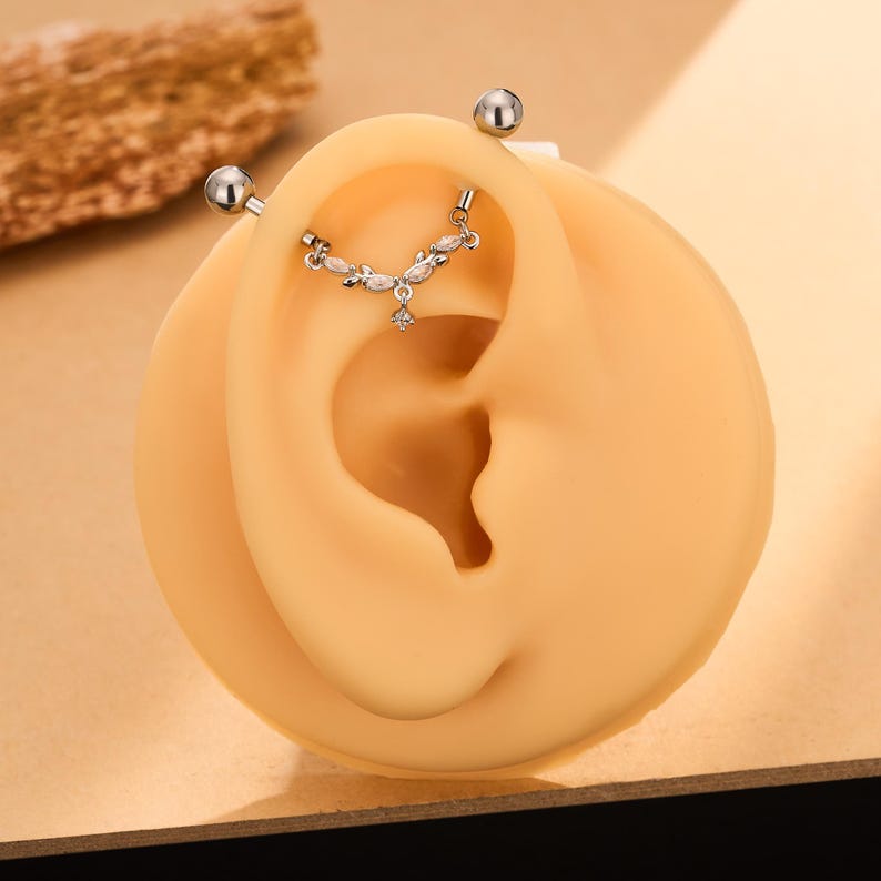 Op de afbeelding: Een zilverkleurige industri&euml;le barbell piercing met een gebogen, bladvormige bedel versierd met kleine, heldere marquise-geslepen stenen en een enkele bungelende kristal, gepresenteerd op een beige siliconen oormodel voor demonstratie.