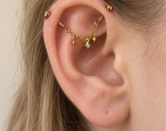 Op de afbeelding: Een gouden industriële barbell-piercing met een fijne ketting die tussen twee uiteinden hangt. De ketting is versierd met drie kleine stervormige bedels, waarbij de middelste ster is voorzien van een minuscuul helder edelsteentje in het midden.