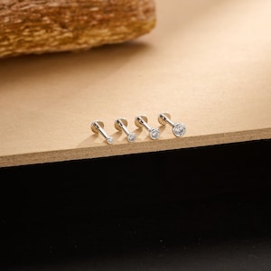 May include: Four silver-colored, flat-backed stud earrings with clear, round gemstones of varying sizes. The earrings are arranged in a row on a light brown surface, with a blurred wooden element in the background.
