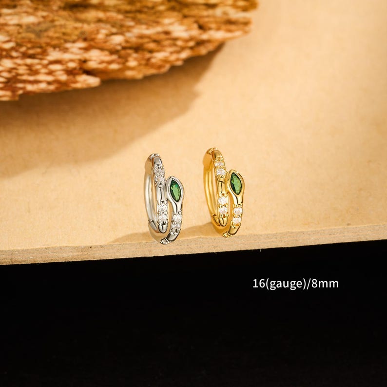 May include: Two hoop earrings, one silver and one gold, each featuring a snake design. The snake's head is adorned with a green gemstone, and the body is embellished with small, clear stones. The text "16(gauge)/8mm" is visible.