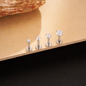 May include: Four silver-colored lip piercing studs with clear, round cubic zirconia stones. The studs are arranged in a row, with the stones increasing in size. The posts are straight with a flat, circular base.