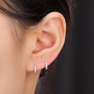 May include: Close-up of an ear with two small silver hoop earrings. Each earring is set with small, clear gemstones. The earrings are shown against a dark background.