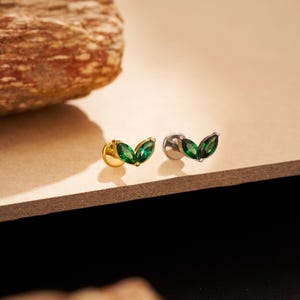 Op de afbeelding: Twee blad-vormige oorstekers. Eén oorbel heeft een gouden pin en achterkant, de andere heeft een zilveren pin en achterkant. Beide oorbellen hebben twee groene, marquise-geslepen edelstenen, gerangschikt om op bladeren te lijken.