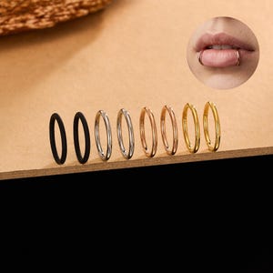 May include: An assortment of seven hoop earrings in various metallic finishes: black, silver, rose gold, and gold. The earrings are arranged in a row on a wooden surface. A close-up of a lip piercing with a gold hoop earring is in the upper right corner.