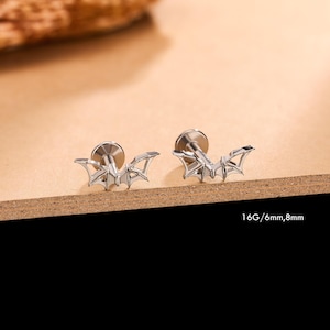 May include: Two silver-toned bat-shaped stud earrings with flat back posts. Each earring features a detailed bat silhouette with spread wings. The text 16G/6mm, 8mm is printed in the lower right corner, indicating the gauge and post lengths.
