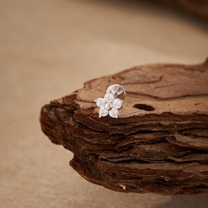 Puede incluir: Un pendiente plateado con forma de flor con piedras claras y brillantes. El pendiente tiene una parte trasera plana y un poste para perforar. El diseño floral tiene cinco pétalos alrededor de una piedra central. El pendiente se muestra sobre madera texturizada.