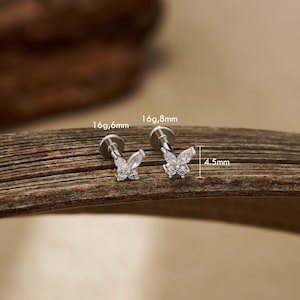 May include: Two silver-colored butterfly-shaped ear studs with clear, sparkling stones. The studs are shown on a wooden surface. One stud is 6.6mm wide, and the other is 8mm wide. The butterfly design is 4.5mm high.
