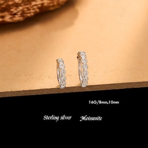 May include: Two sterling silver hoop earrings featuring a row of bezel-set moissanite stones. The text 16G/8mm, 10mm, Sterling silver, and Moissanite is printed below the earrings, which are displayed in two different sizes.