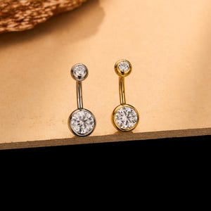 May include: Two curved barbell body jewelry pieces, one in silver-tone metal and one in gold-tone metal. Each piece features a small round clear gemstone at the top and a larger round clear gemstone at the bottom, both set in a bezel setting.