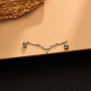 Op de afbeelding: Een zilverkleurige tepelpiercing met een V-vormig decoratief middenstuk versierd met kleine doorzichtige steentjes. Het sieraad bevat twee rechte staafjes met schroefdraad en twee gepolijste bolletjes aan weerszijden van het centrale ontwerp.