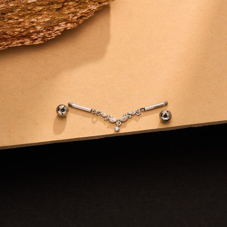 Op de afbeelding: Een zilverkleurige tepelpiercing met een V-vormig decoratief middenstuk versierd met kleine doorzichtige steentjes. Het sieraad bevat twee rechte staafjes met schroefdraad en twee gepolijste bolletjes aan weerszijden van het centrale ontwerp.