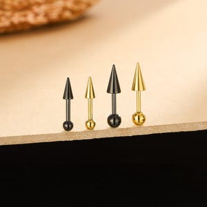 May include: Four ear piercing studs with cone-shaped tops and ball ends. Two are black, and two are gold-colored. The studs are arranged in a row on a light brown surface, with a dark background.