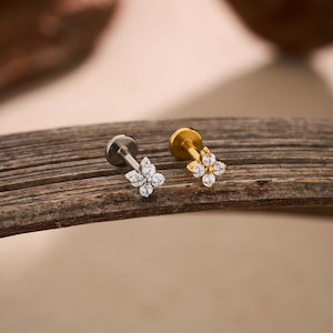 May include: Two flower-shaped ear studs. One is silver-colored, the other is gold-colored. Each stud features four clear, round gemstones. The studs are displayed on a wooden surface with a neutral background. The design is delicate and suitable for various occasions.
