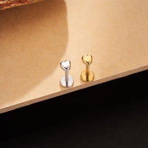 May include: Two lip piercing studs. One is silver-colored, and the other is gold-colored. Each stud features a heart-shaped top and a flat base. The studs are displayed on a light brown surface.