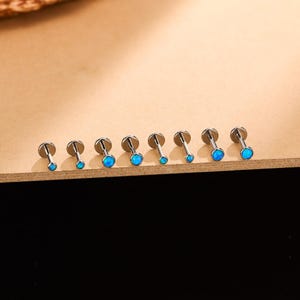May include: A collection of seven silver-colored lip piercing studs with blue opal-like stones. The studs are arranged in a row on a light brown surface, with a dark background. The stones vary slightly in size.