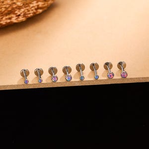 Puede incluir: Una colección de siete piercings labret plateados con parte trasera plana y piedras iridiscentes tipo ópalo. Las piedras varían en tamaño y color, desde rosa hasta azul. Los piercings están dispuestos en fila sobre una superficie marrón claro.