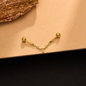 Op de afbeelding: Een goudkleurige industri&euml;le barbell piercing met een centrale V-vormige hanger. De hanger is versierd met kleine, heldere, marquise-geslepen stenen en een enkele ronde steen aan het onderste puntje, verbonden met twee gladde bolvormige uiteinden.
