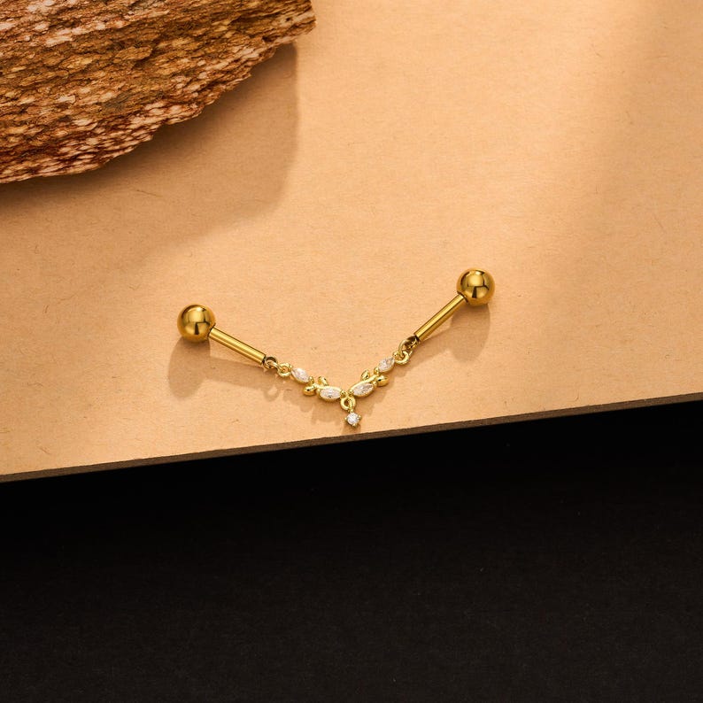 Op de afbeelding: Een goudkleurige industri&euml;le barbell piercing met een centrale V-vormige hanger. De hanger is versierd met kleine, heldere, marquise-geslepen stenen en een enkele ronde steen aan het onderste puntje, verbonden met twee gladde bolvormige uiteinden.