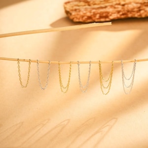 May include: A selection of chain earrings in gold and silver hues. The earrings are displayed on a thin wooden rod, set against a warm, neutral backdrop. Some earrings have a single chain, while others feature a double chain design.